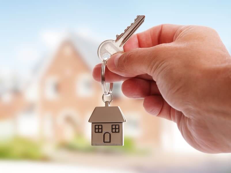 A Hand Holding House Keys On House Shaped Keychain In Front Of A New Home