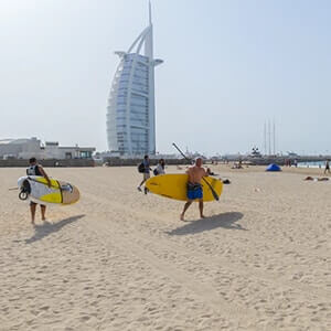 People walking across a sandy beach to the sea with Sharjah buildings in the background