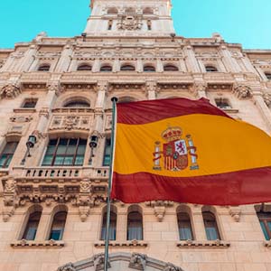 A Spanish flag with a tall Madrid building in the background, in a clear blue-sky day