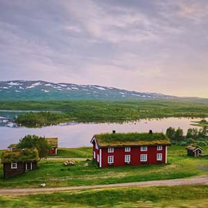 Red wooden houses with grassy roofs next to a lake set in green Swedish countryside and a mountain in the background