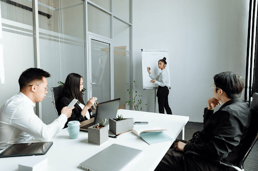 Woman writing on whiteboard and 3 others brainstorming at work table.
