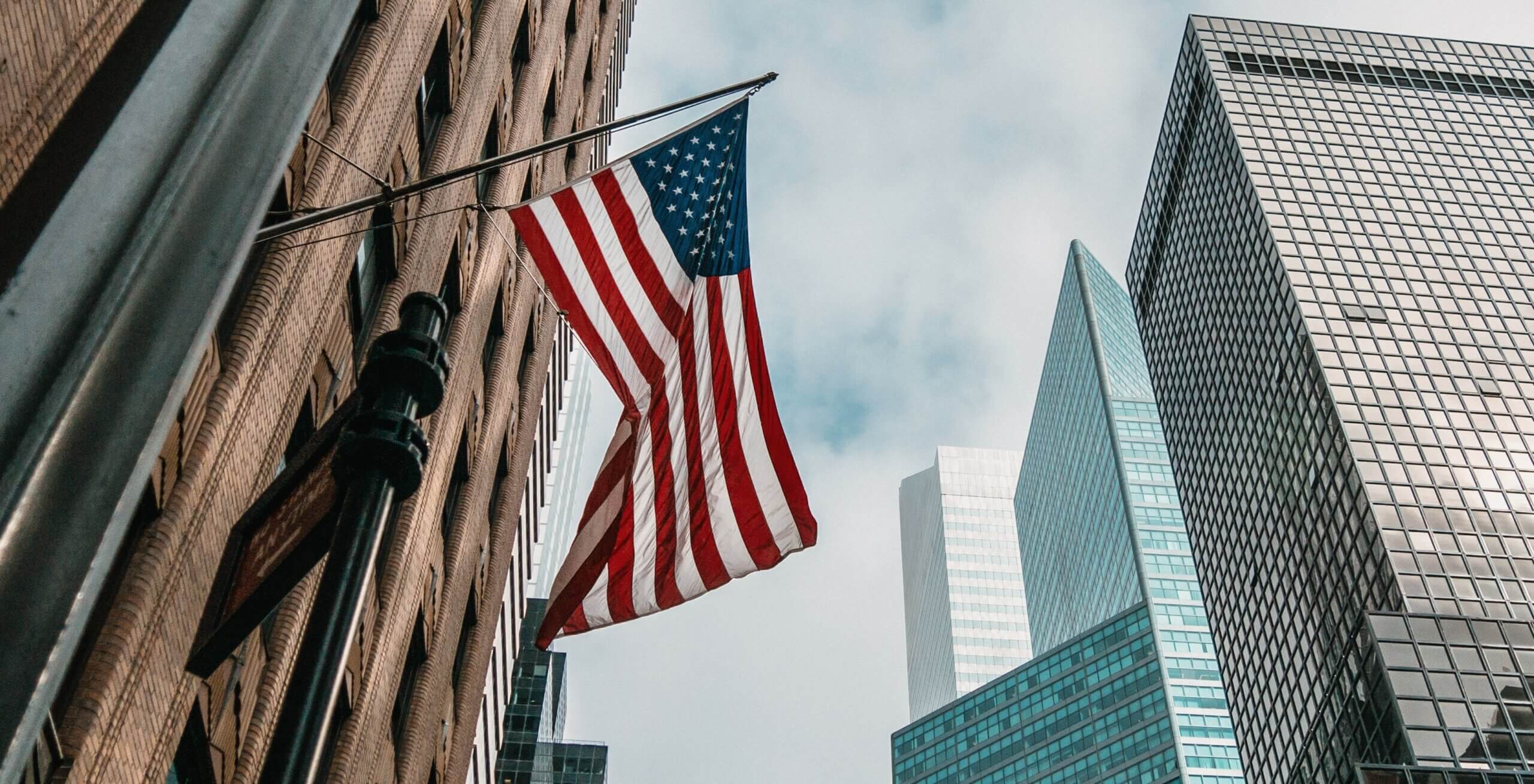 The USA Or United States Of America Flag On A Flagpole Near Skyscrapers Under A Cloudy Sky