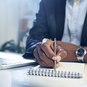 Man in a suit making notes on a pad next to a laptop