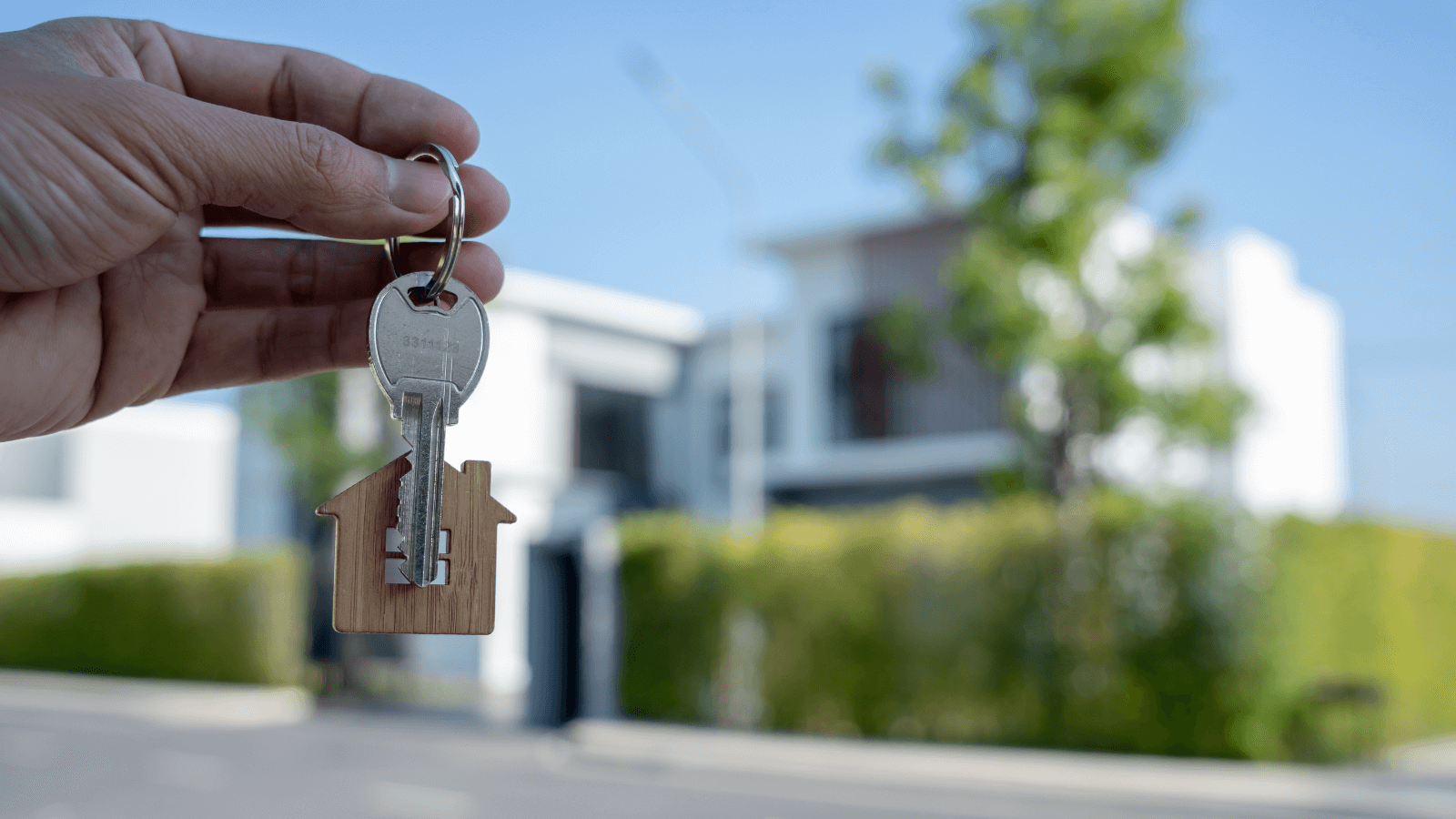 Hand Holding A Set Of House Keys In The Foreground, Blurred House In The Background