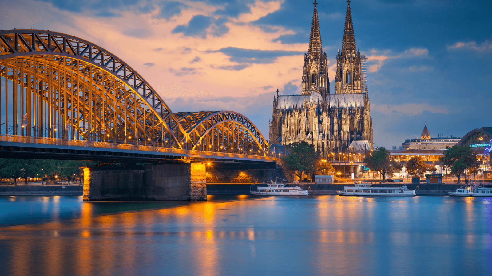 The Cologne Cathedral And Hohenzollern Bridge Across The Rhine River In Germany